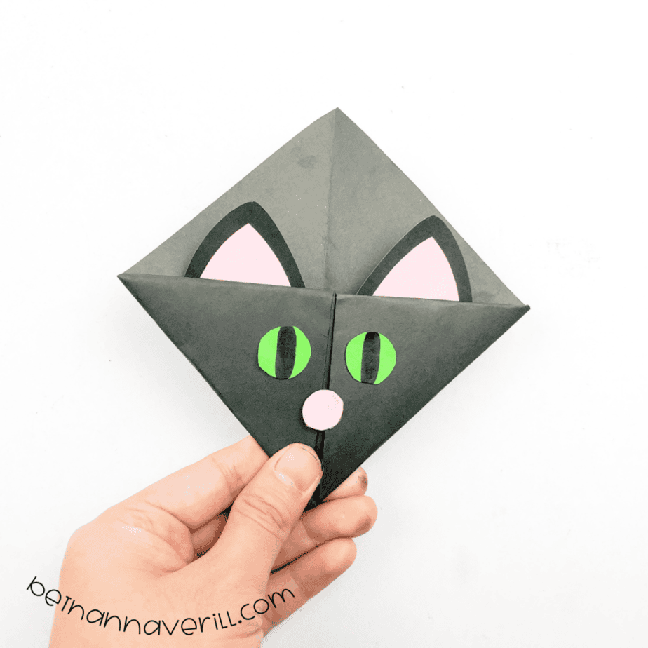 Hand holding a finished black origami cat corner bookmark with green eyes