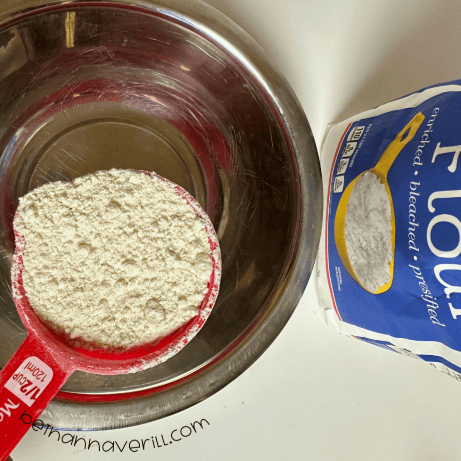 Measuring cup of flour added to a metal mixing bowl for cinnamon applesauce playdough