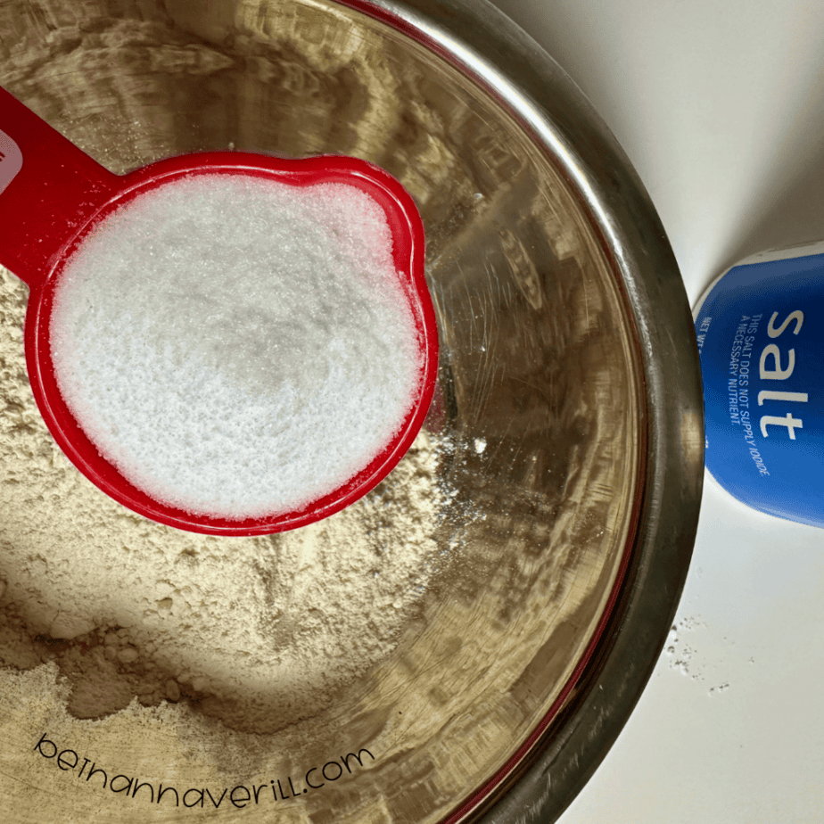 Measuring cup of salt poured over flour in a mixing bowl