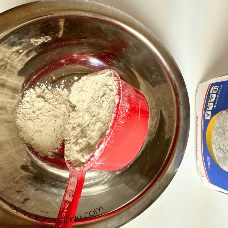Measuring cup of flour added to a metal mixing bowl for apple playdough