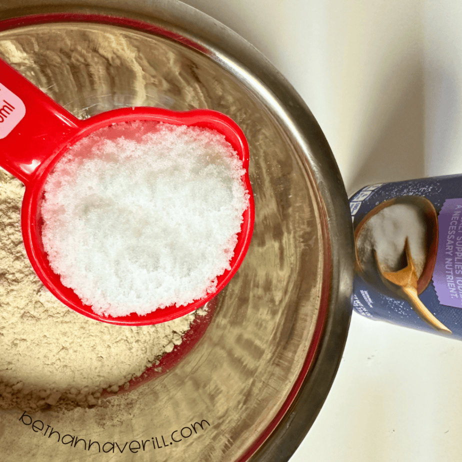Measuring cup of salt poured over flour in a mixing bowl