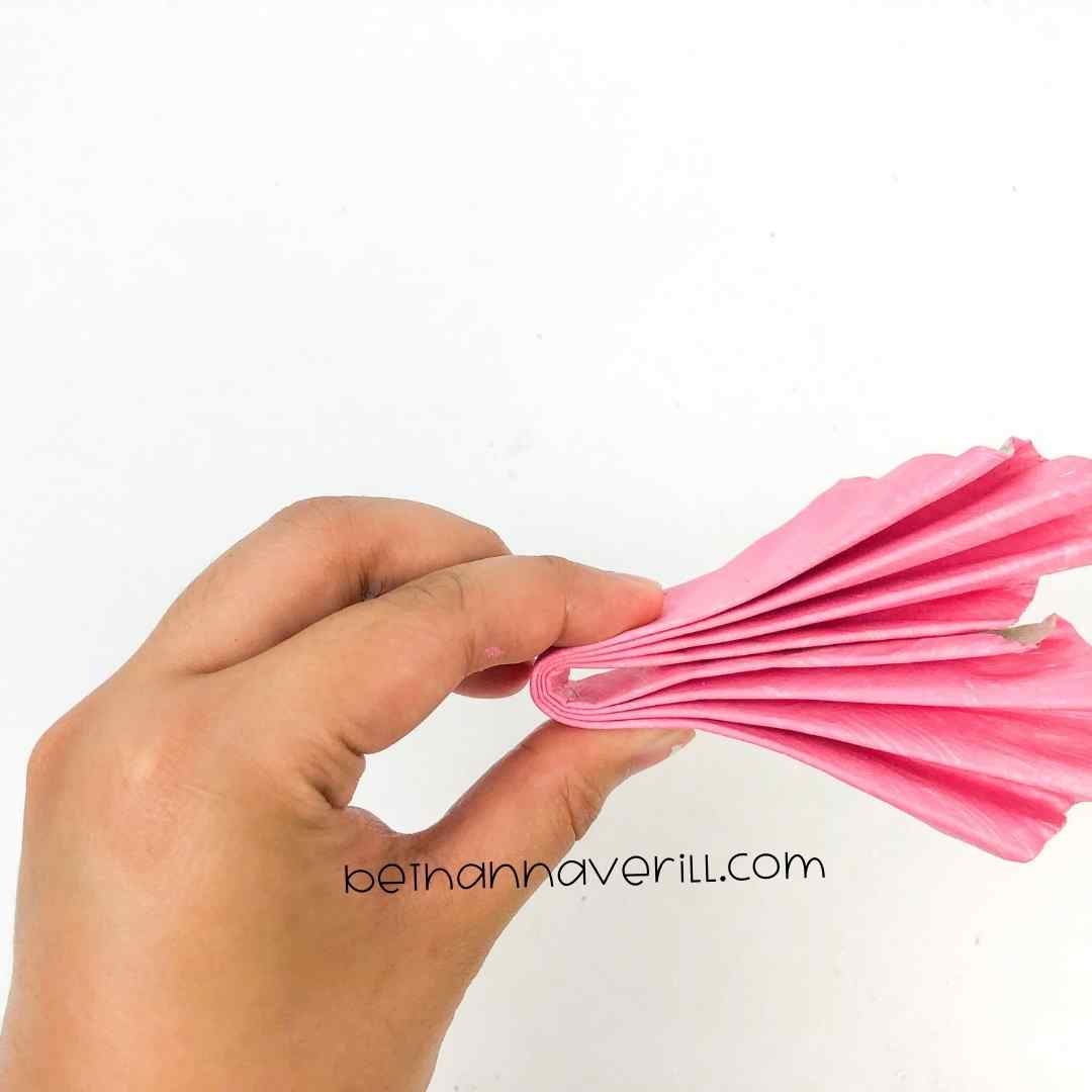 A hand pressing two sides of a folded pink paper plate together with double-sided tape applied for crafting a heart.