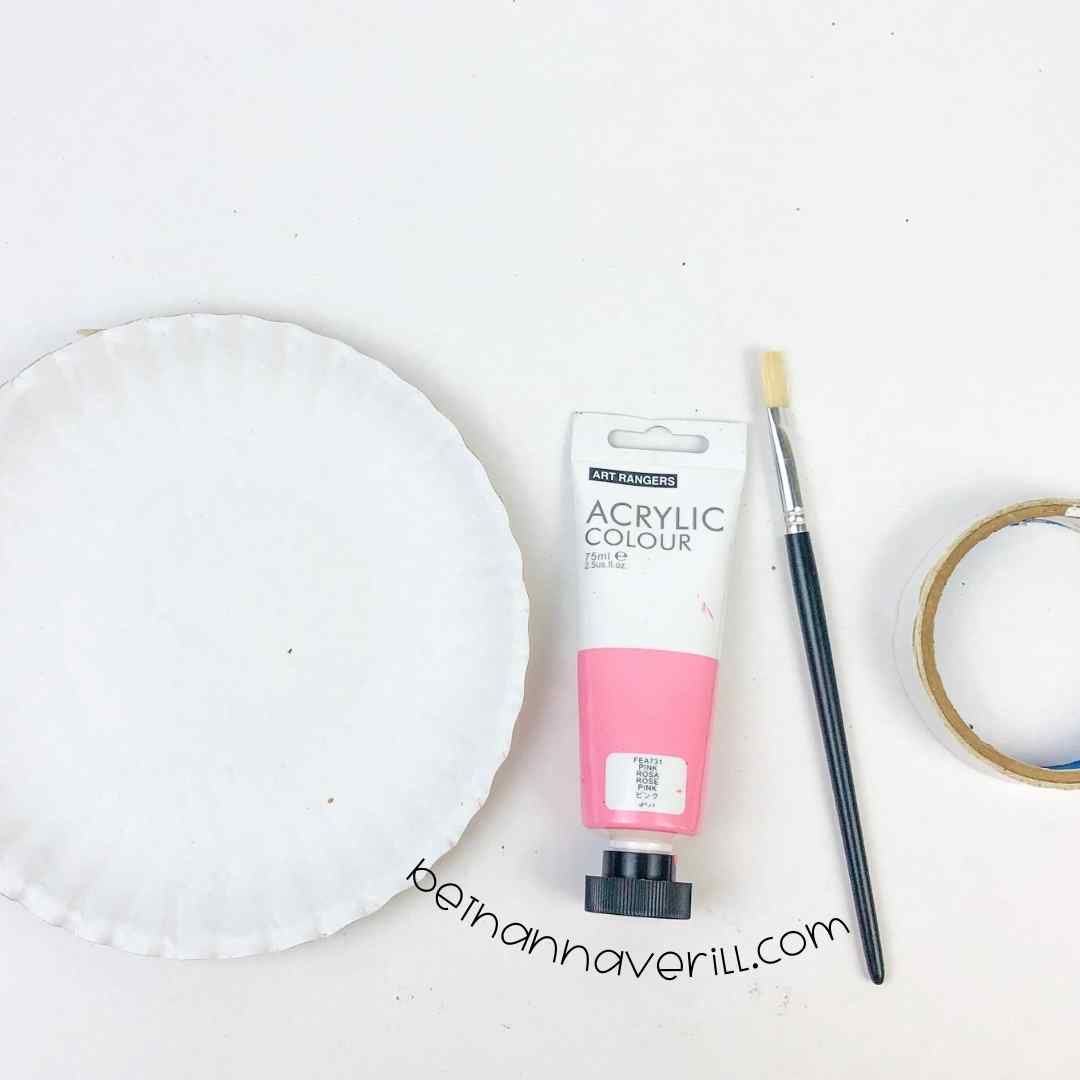 Supplies for a paper plate heart craft: a white paper plate, pink acrylic paint, a paintbrush, and double-sided tape.
