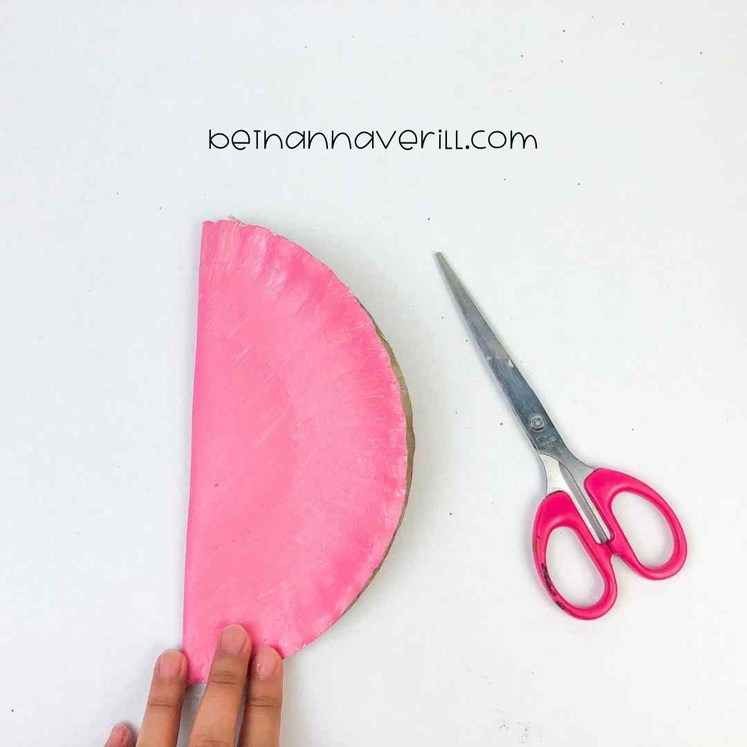 A pink-painted paper plate folded in half with a pair of pink scissors beside it, illustrating the folding step in a DIY craft project.