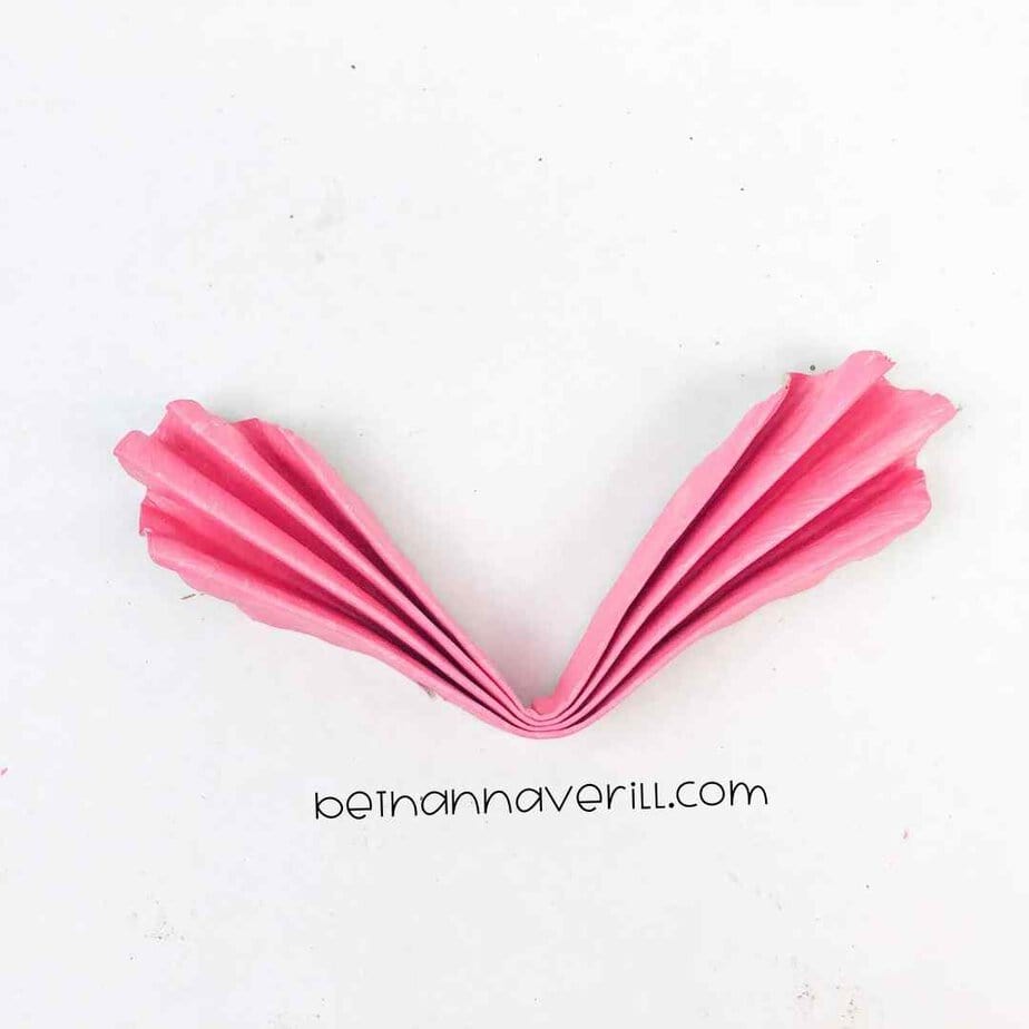 A pink accordion-folded paper plate being bent in half by a hand, showcasing a step in creating a folded heart craft.