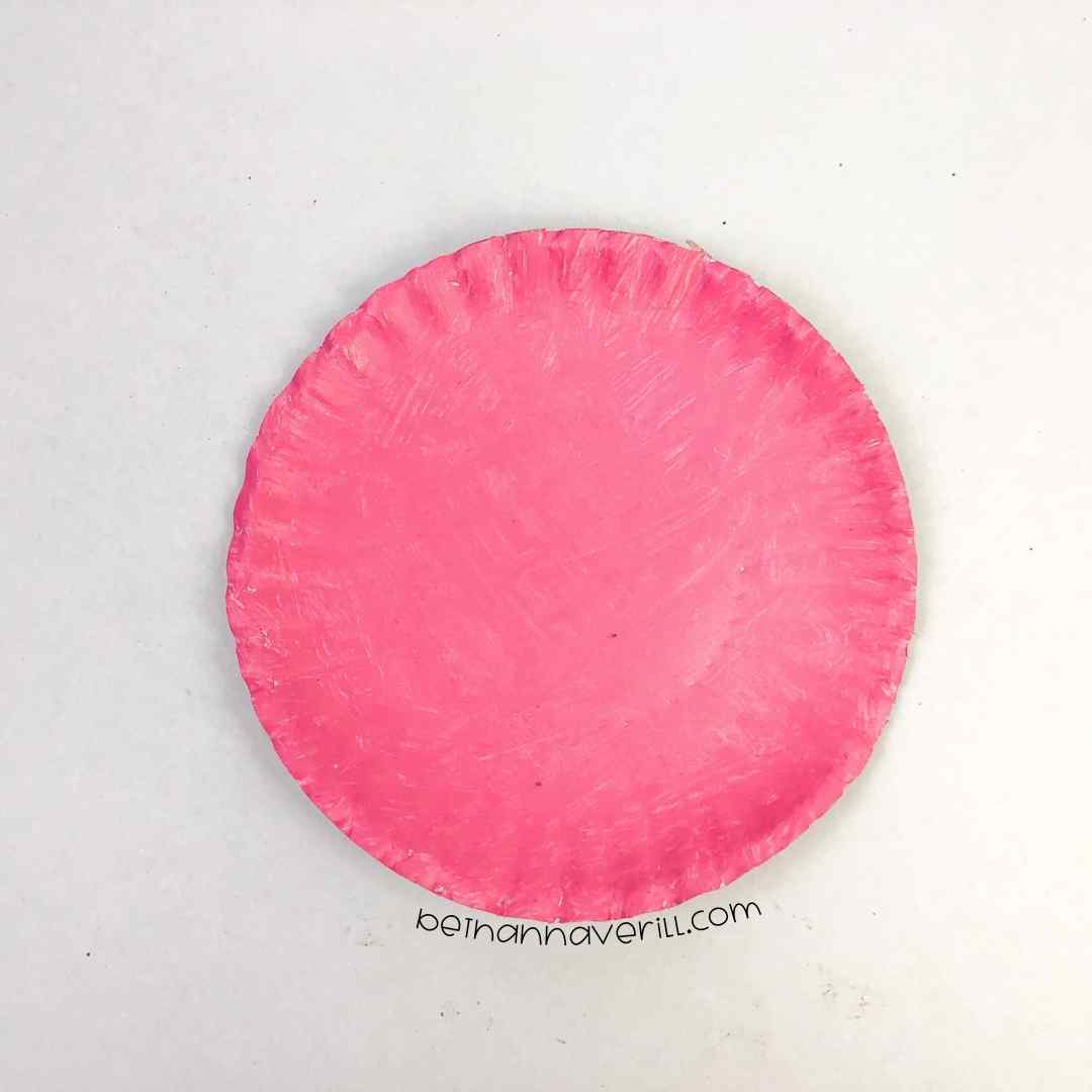 A plain white paper plate painted pink with visible brush strokes, part of a DIY folded heart craft project.