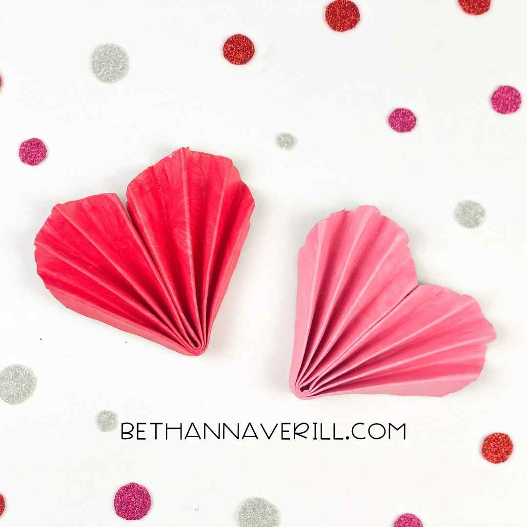 Two paper hearts crafted from folded pink and red paper plates, displayed on a white surface with glittery confetti dots.