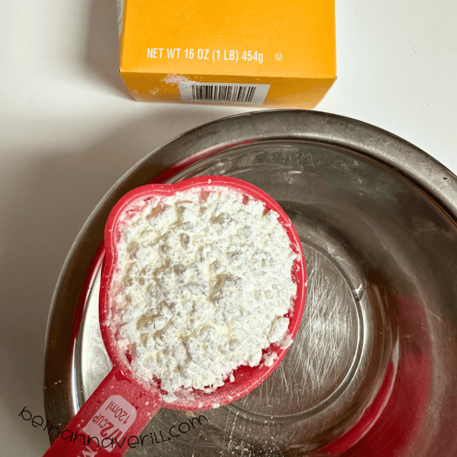 Measuring cup of cornstarch added to a metal mixing bowl for snow dough
