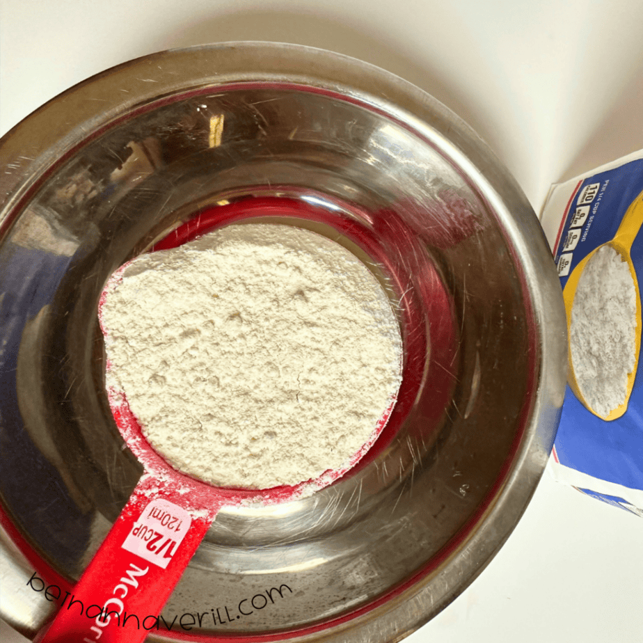 Measuring cup of flour added to a metal mixing bowl for vanilla playdough