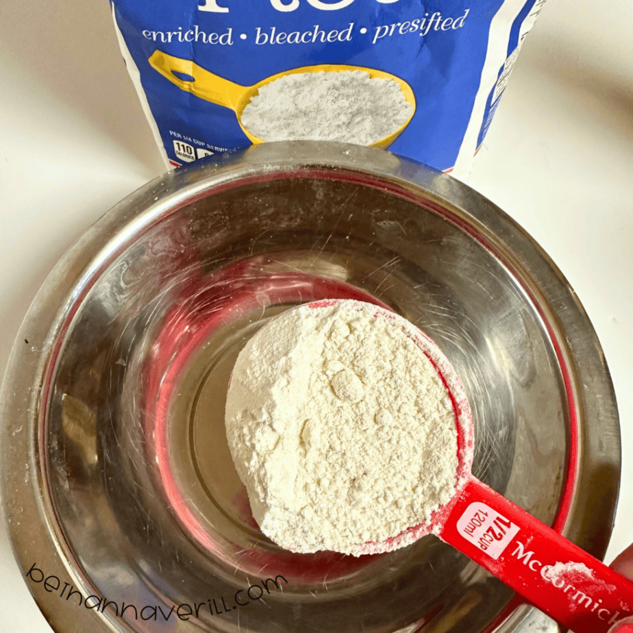Measuring cup of flour added to a metal mixing bowl for hot cocoa playdough