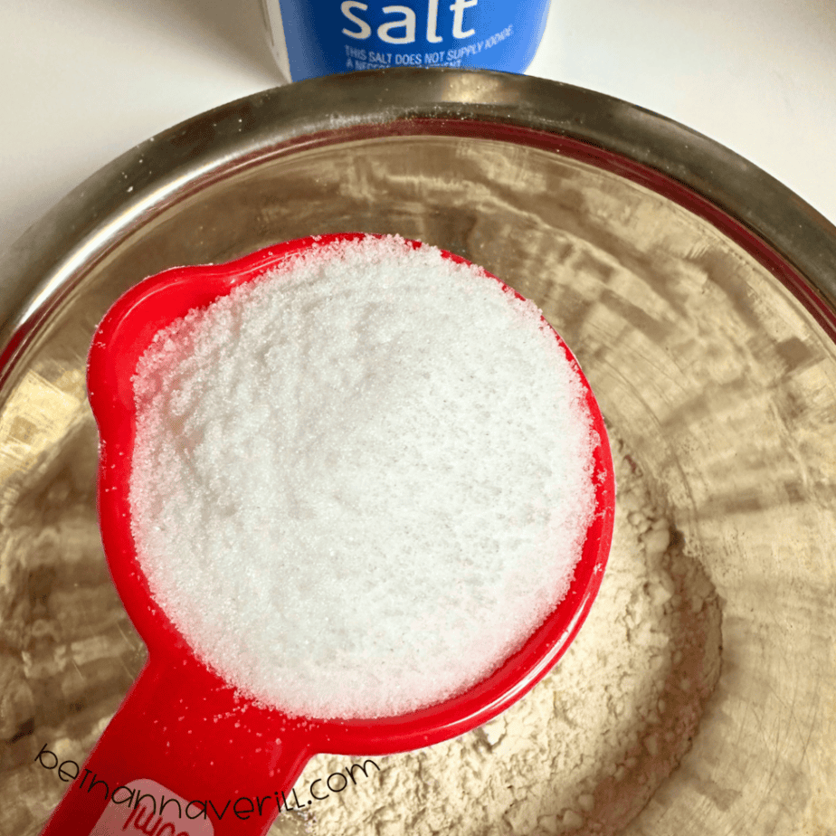 Measuring cup of salt poured over the flour in a mixing bowl