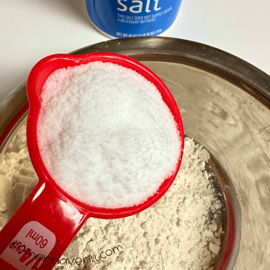 Measuring cup of salt poured over flour in a mixing bowl