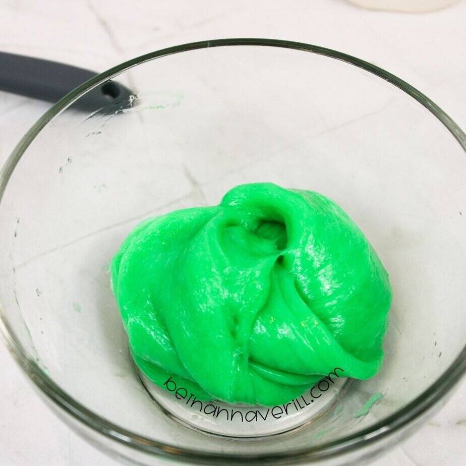 Smooth, shiny green glow-in-the-dark slime formed into a soft mound in a glass bowl, surrounded by minimal ingredients.
