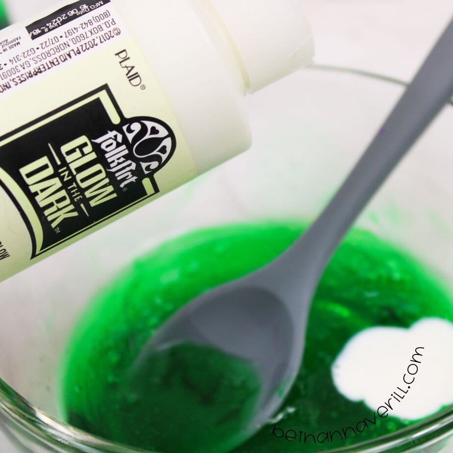A bottle of FolkArt glow-in-the-dark paint is being poured into a glass bowl of green slime base with a gray spoon in view.
