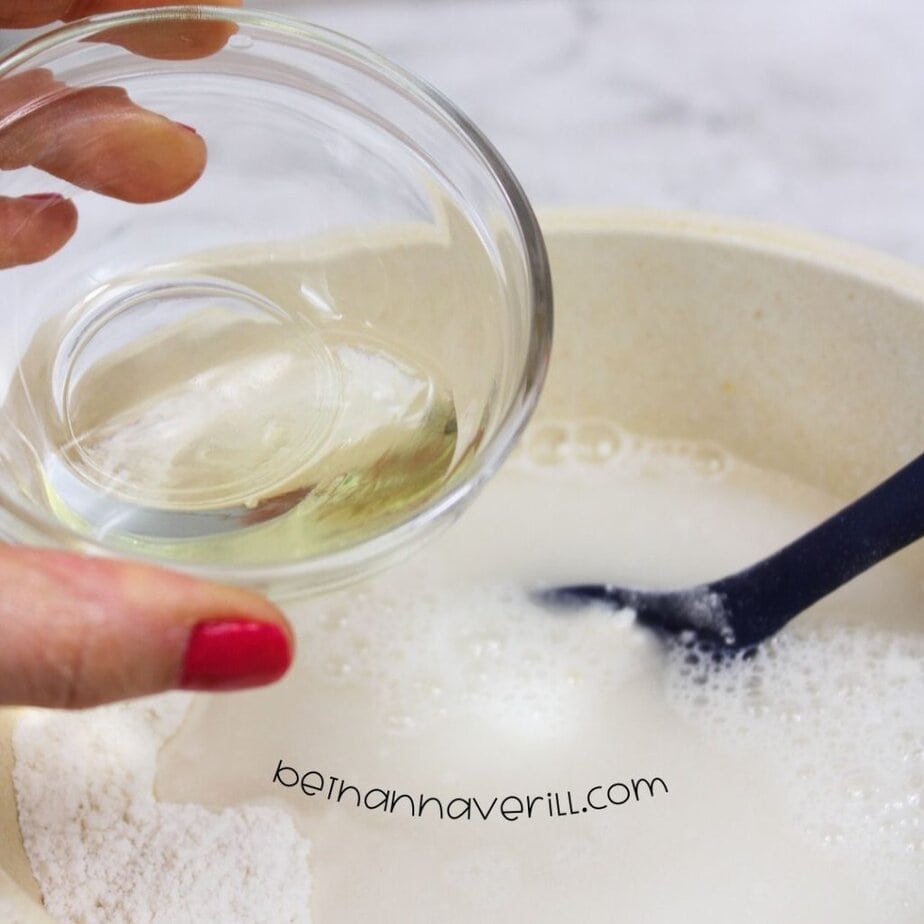 A hand with red-painted nails holds a small glass bowl of oil over a mixing bowl filled with white liquid ingredients and a black spoon submerged in the mixture.