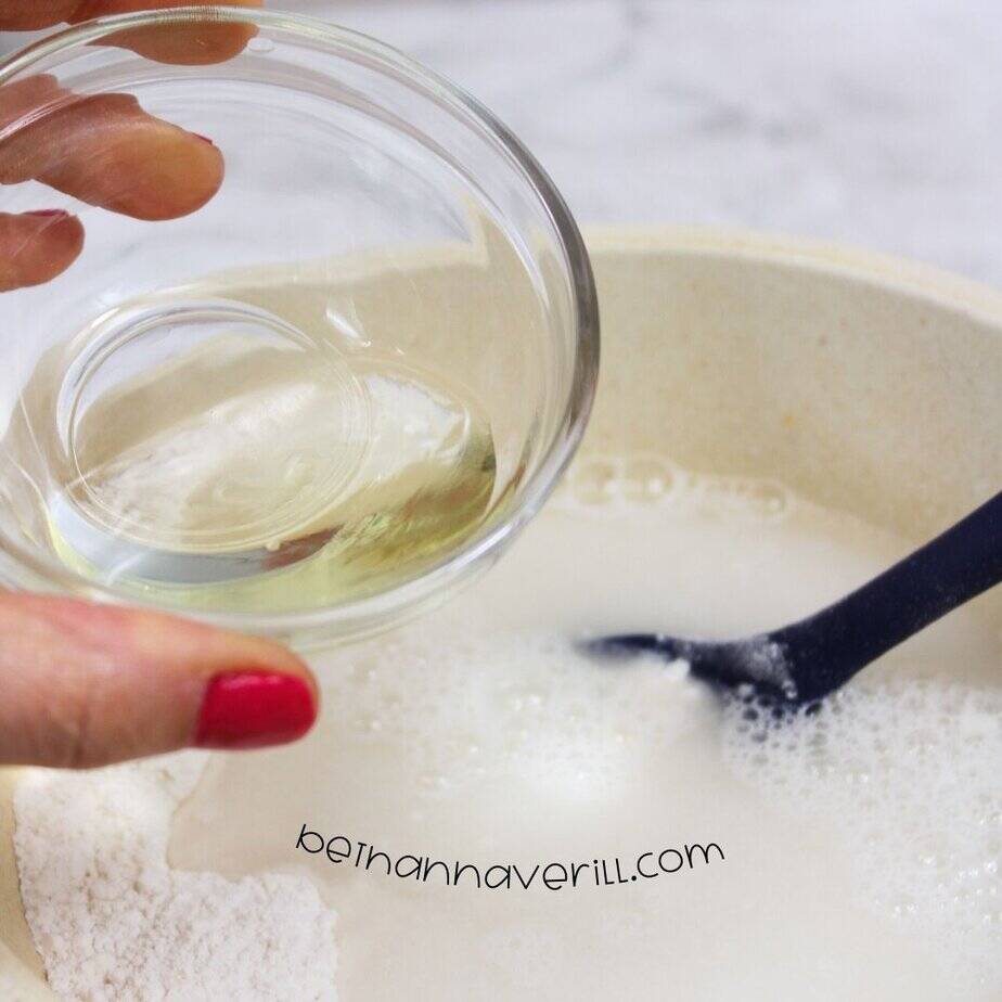 A hand with red-painted nails holds a small glass bowl of oil over a mixing bowl filled with white liquid ingredients and a black spoon submerged in the mixture.