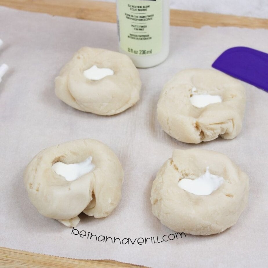 Four beige dough balls rest on parchment paper, each with a well in the center filled with a small amount of glow-in-the-dark paint; a paint bottle and spatula sit nearby.