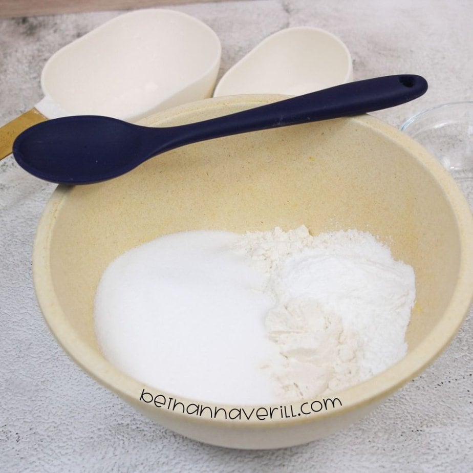 A beige mixing bowl contains white sugar, flour, and a dry powder, with a navy blue spoon resting across the top; two measuring scoops sit in the background.