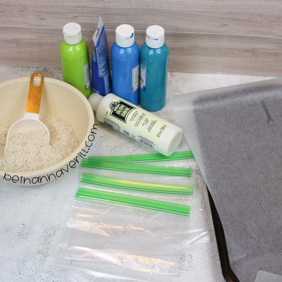 A flat lay of ingredients and supplies including dry rice in a bowl, neon and blue paints, glow-in-the-dark paint, zip-top sandwich bags, and a baking sheet with parchment paper.