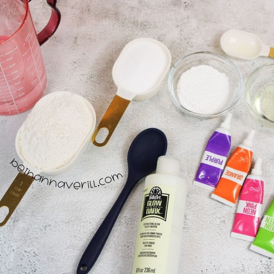 All ingredients and tools laid out on a countertop: measuring cups of flour, sugar, cornstarch, glow-in-the-dark paint, food coloring tubes in purple, orange, neon pink, neon green, a spoon, and a measuring cup.