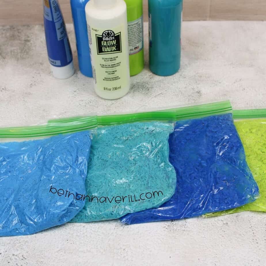 Four sealed zip-top bags filled with dyed rice in shades of light blue, teal, royal blue, and neon green are lined up in front of corresponding paint bottles.