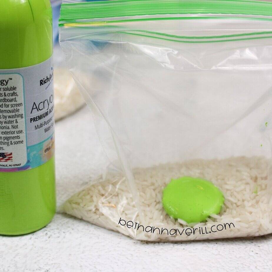 A close-up of a sandwich bag filled with rice and a bright green blob of acrylic paint sitting on top, next to a bottle of green paint.