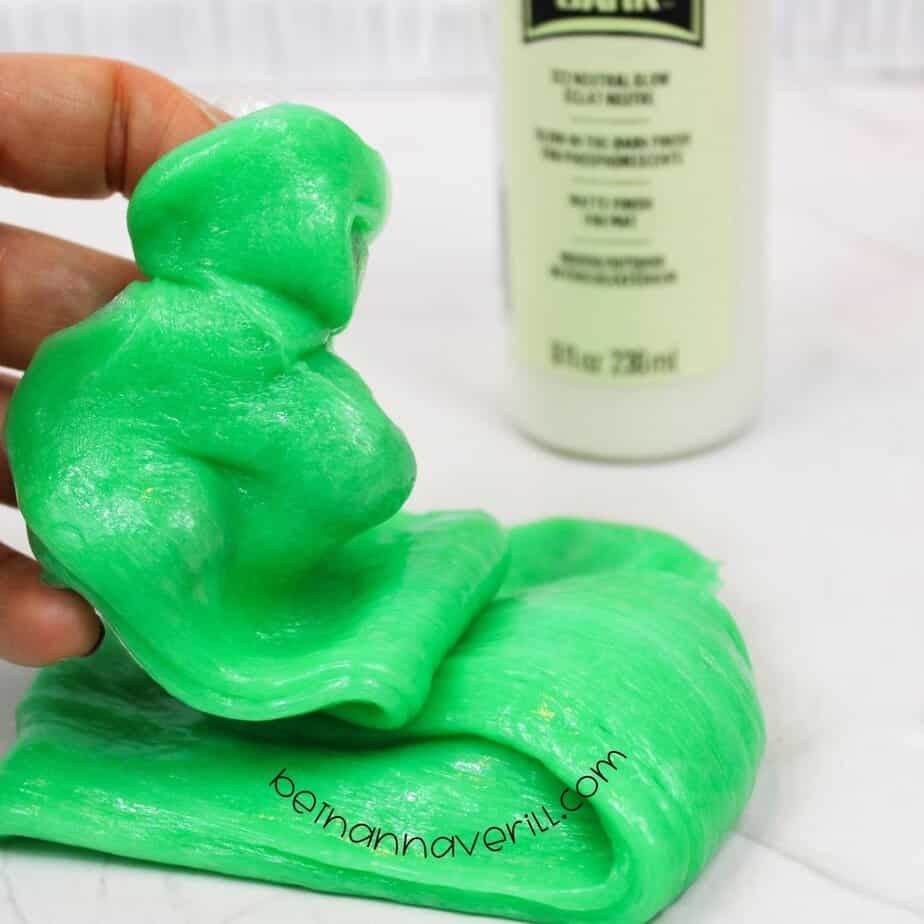A hand stretches a lump of neon green glow-in-the-dark slime, with a bottle of FolkArt glow paint in the background. The slime is glossy, stretchy, and vibrant.