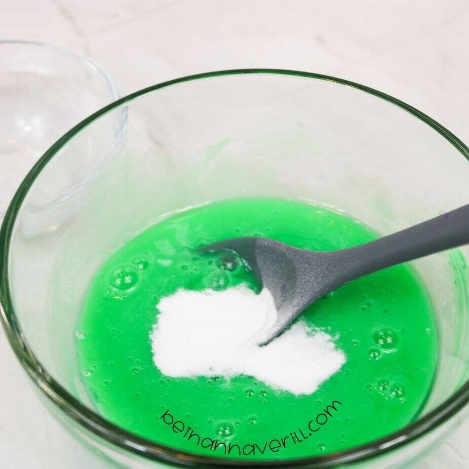 A clear mixing bowl filled with bright green slime mixture, with a pile of baking soda in the center and a gray spoon resting inside.
