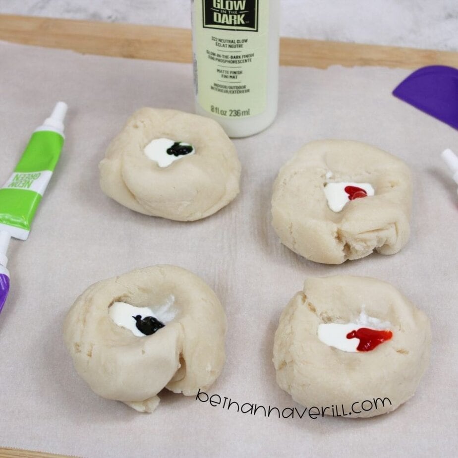 Four plain dough balls with small amounts of glow paint and food coloring (black and red) added to their centers, sitting on parchment paper beside paint supplies.
