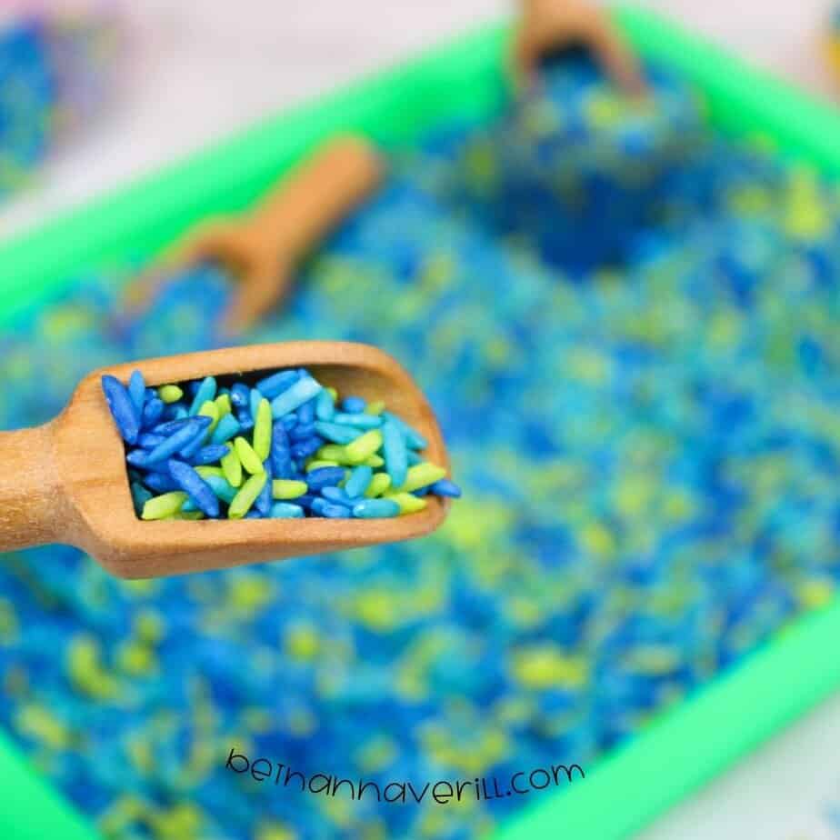 Close-up of a wooden scoop holding bright neon green and blue dyed rice, with a green sensory bin full of rice blurred in the background.