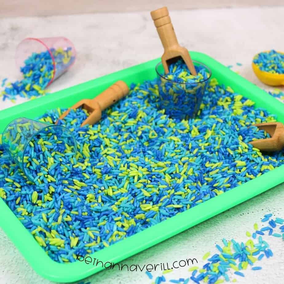 A green plastic bin filled with a mix of dried blue and neon green rice, surrounded by small scoops and cups for play.