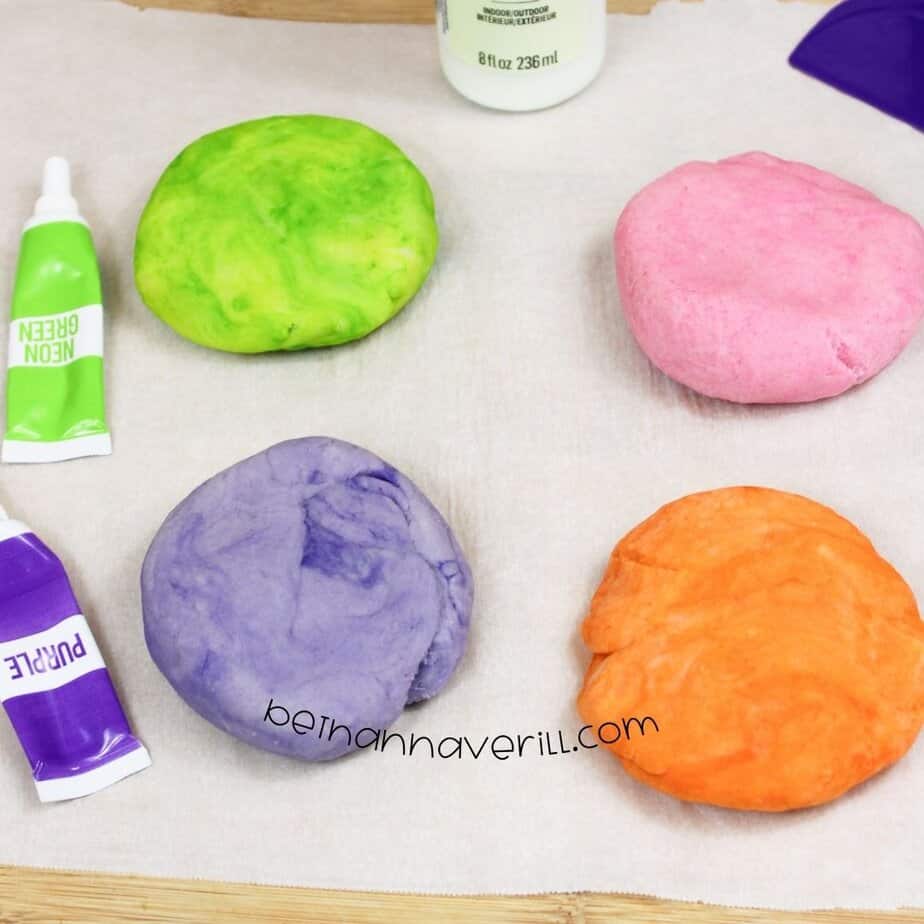 Four flattened discs of play dough—green, pink, orange, and purple—sit on parchment paper next to tubes of corresponding food coloring and a glow paint bottle.