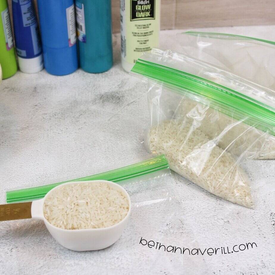 Measuring cup of dry rice sits in the foreground, with clear plastic sandwich bags filled partially with rice, glow paint, and blue paint bottles in the background.