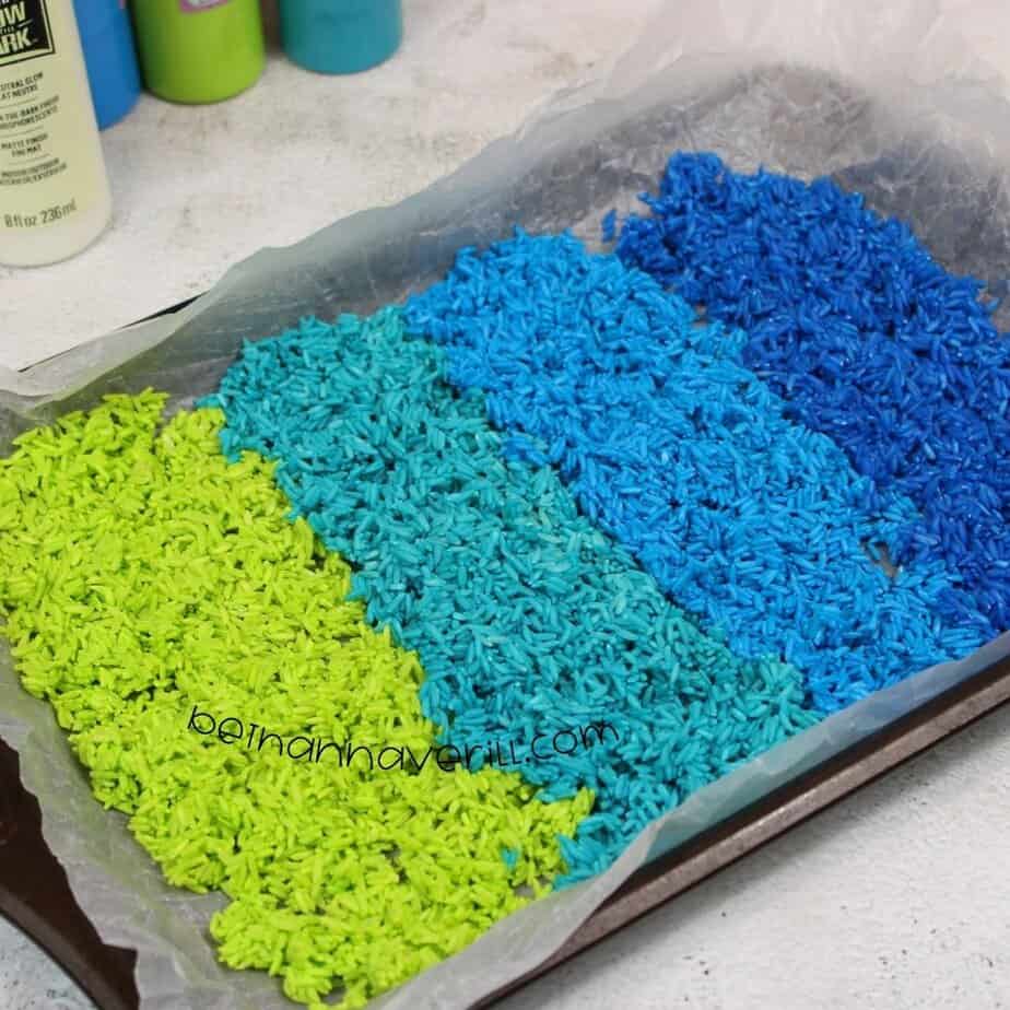 A baking sheet lined with parchment paper displays neat rows of dyed rice—neon green, teal, light blue, and dark blue—ready to dry.