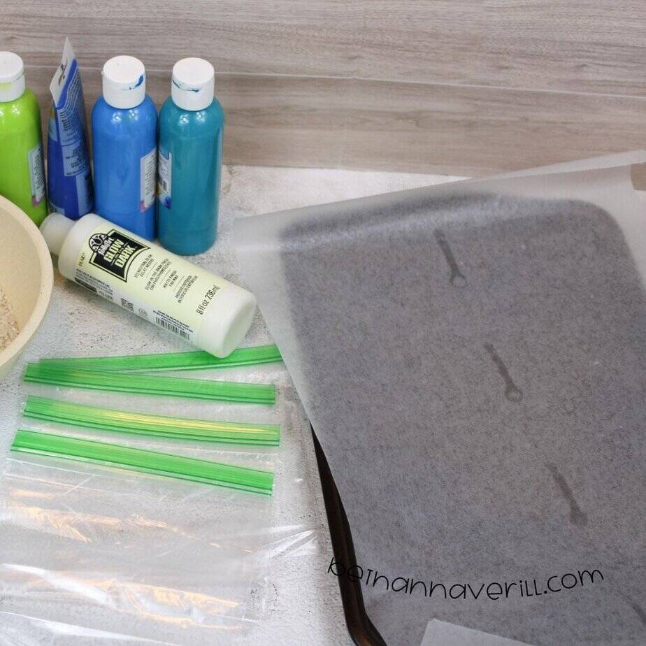 A baking sheet covered with parchment paper sits beside a cluster of paints, glow-in-the-dark paint, and sandwich bags on a textured surface.