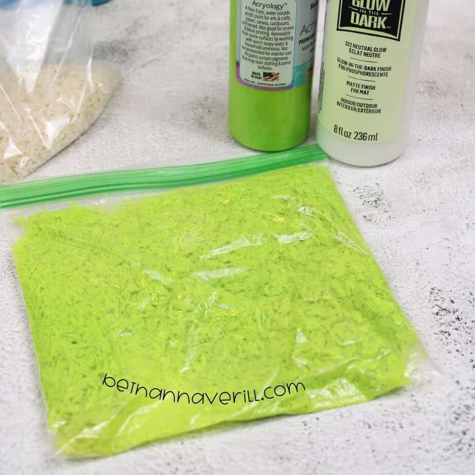 A zip-top bag filled with bright neon green rice sits sealed on the counter, with bottles of green and glow-in-the-dark paint in the background.