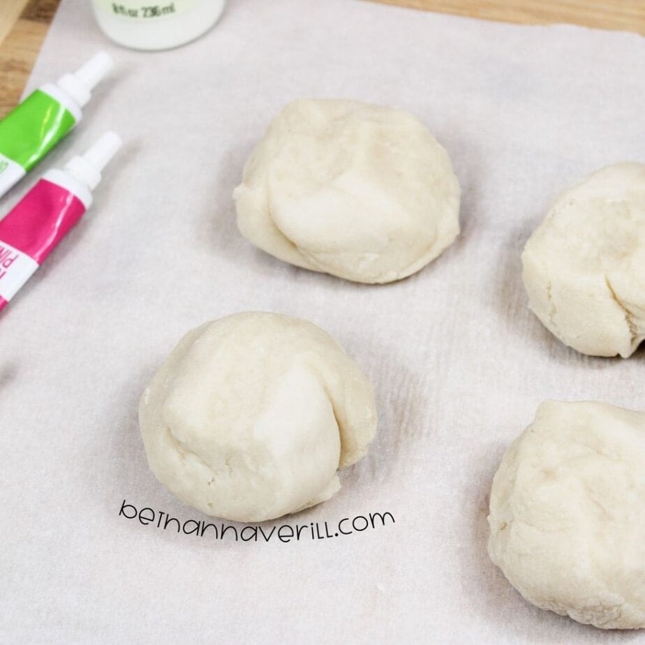 Four uncolored dough balls on parchment paper, positioned near tubes of neon green and neon pink food coloring and a glow-in-the-dark paint bottle.