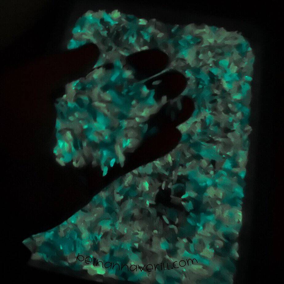 The sensory bin glows in the dark, with a hand scooping up teal-glowing rice above a full tray, creating a magical effect.