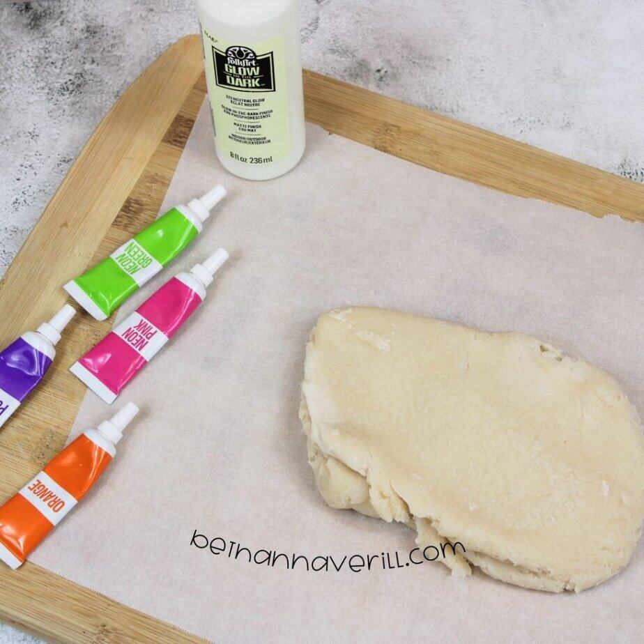 A single slab of cooked dough rests on parchment paper next to tubes of colorful food dye and a glow paint bottle, ready to be divided and colored.