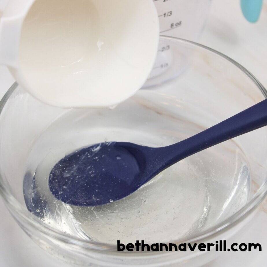 A white liquid, likely glue or contact solution, is being poured into a bowl of clear slime mixture, with a navy spoon resting inside.