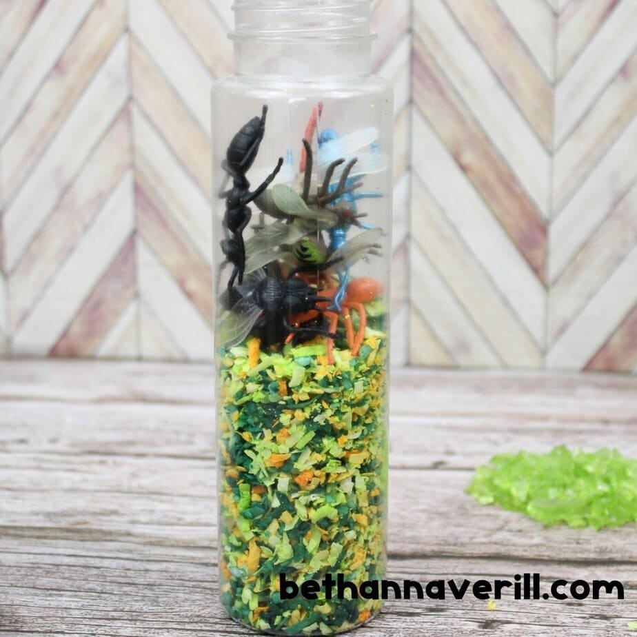 A clear bottle filled halfway with shredded coconut has a pile of toy bugs sitting on top, ready to be added in. The colorful insects include spiders, ants, and dragonflies.