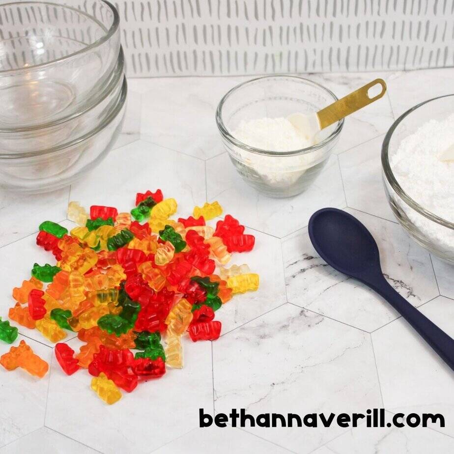 A flat lay image showing ingredients to make edible gummy bear slime: a colorful pile of gummy bears, bowls of white powder (likely cornstarch or powdered sugar), a navy spoon, and clear mixing bowls. The surface is white with a faint hexagonal pattern, and the website “bethannaverill.com” is displayed in the bottom right corner.
