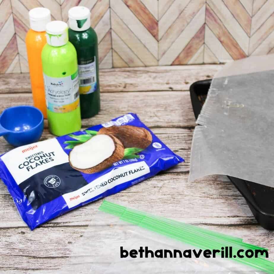 A package of Meijer sweetened coconut flakes, green and yellow acrylic paints, a blue scoop, and wax paper are laid out on a rustic wood surface, everything needed to start the project.