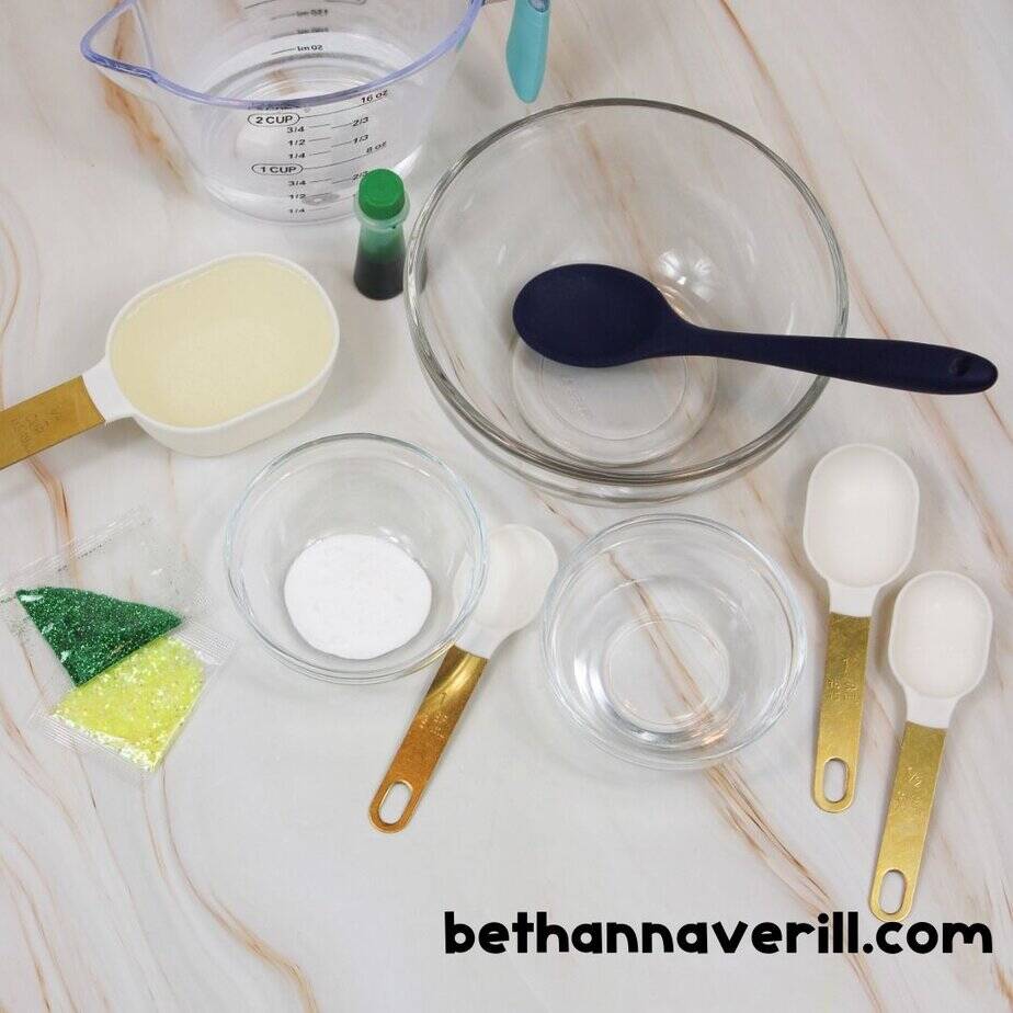 A flat lay of ingredients including glue, baking soda, glitter, green dye, measuring spoons, bowls, and a spoon, neatly arranged on a white marble surface.