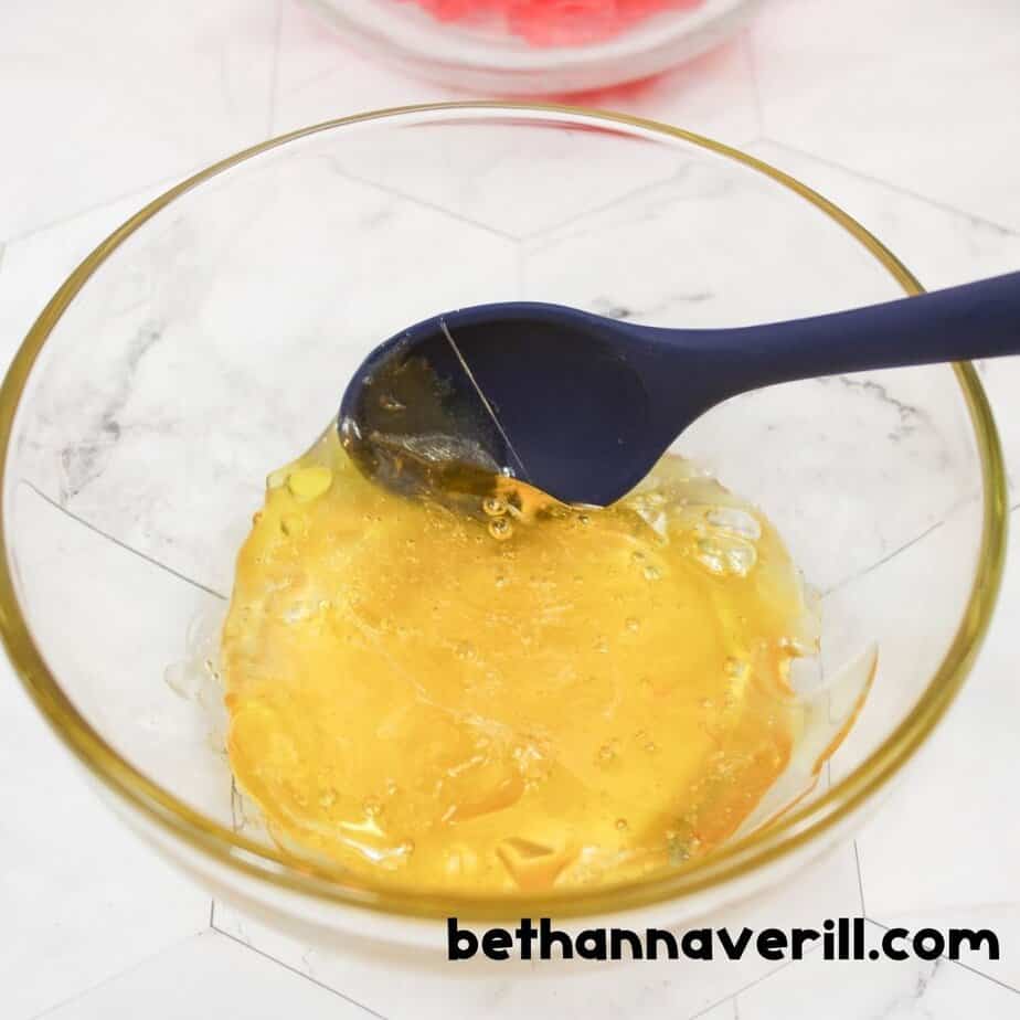A glass bowl holds melted yellow gummy bears with a navy spoon resting inside, showing the gooey texture of the candy after heating. Website link is placed at the bottom.