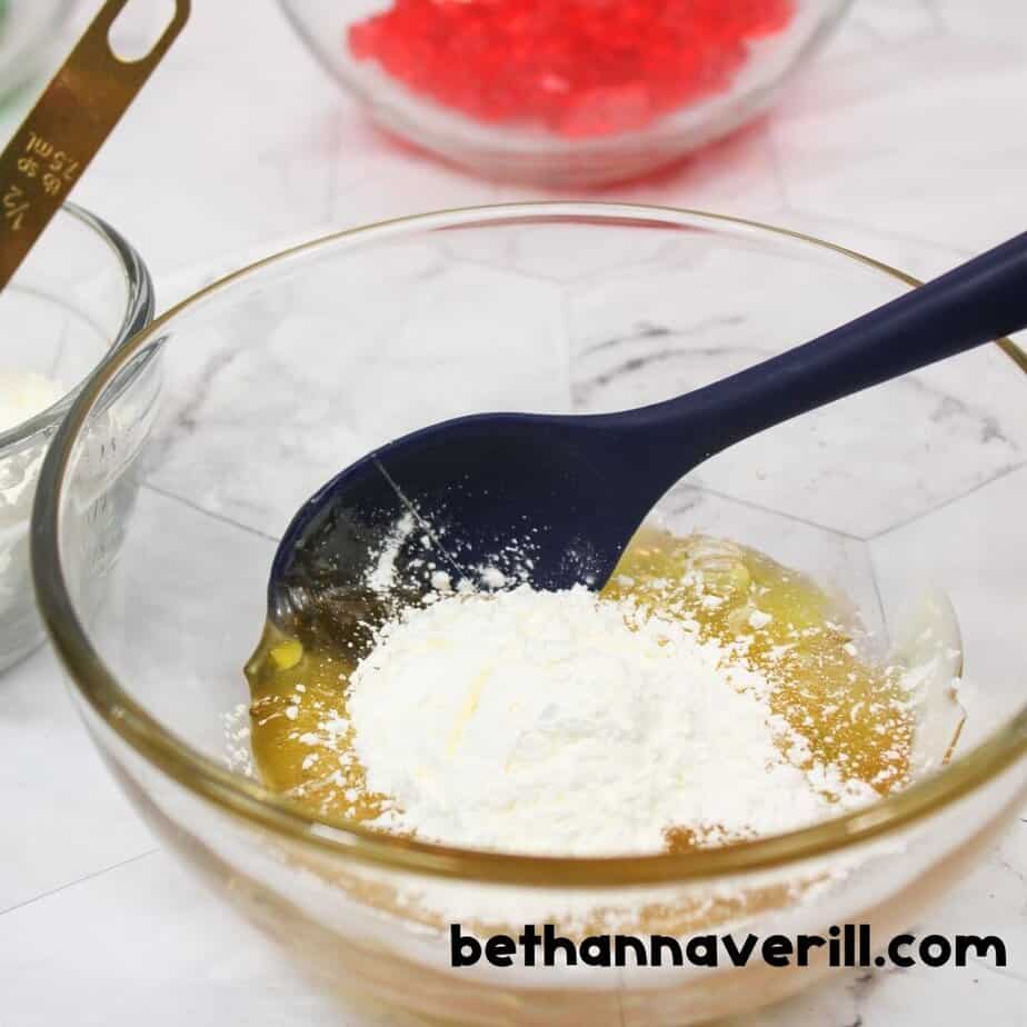 Cornstarch is being stirred into melted gummy bears in a glass bowl using a navy spoon. The white powder is mid-mix, sitting atop the sticky yellow mixture.