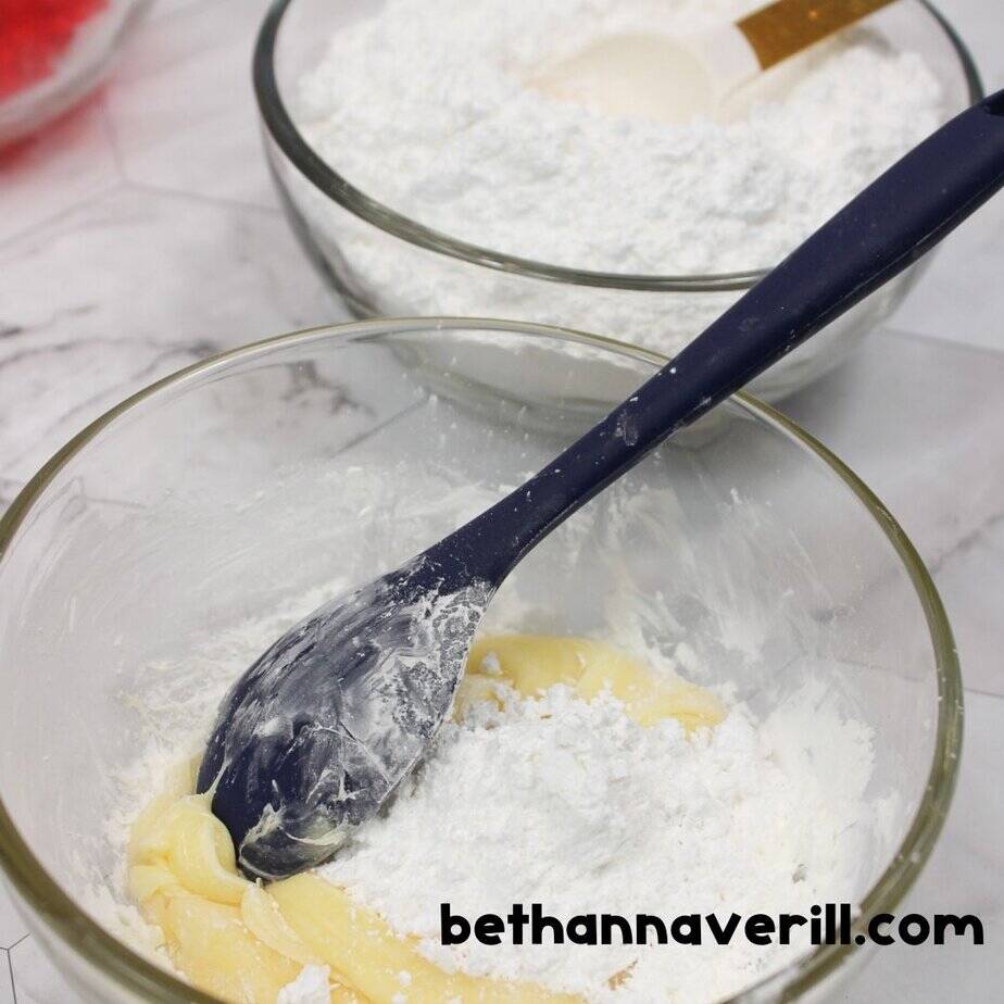 A glass bowl filled with thick yellow melted gummy bears topped with powdered sugar, being mixed with a navy spoon. A second bowl of powdered sugar sits behind it.