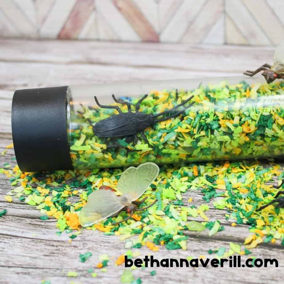 A sensory bottle lies on its side, filled with colorful shredded coconut and plastic insects like a beetle and a winged bug. Some filler spills out onto the wooden surface.