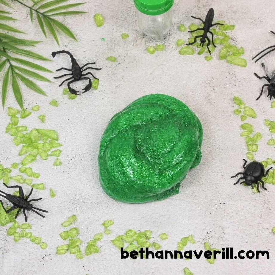 A swirled lump of green glitter slime is placed in the center of a bug-themed scene with toy scorpions, beetles, and faux greenery for dramatic effect.