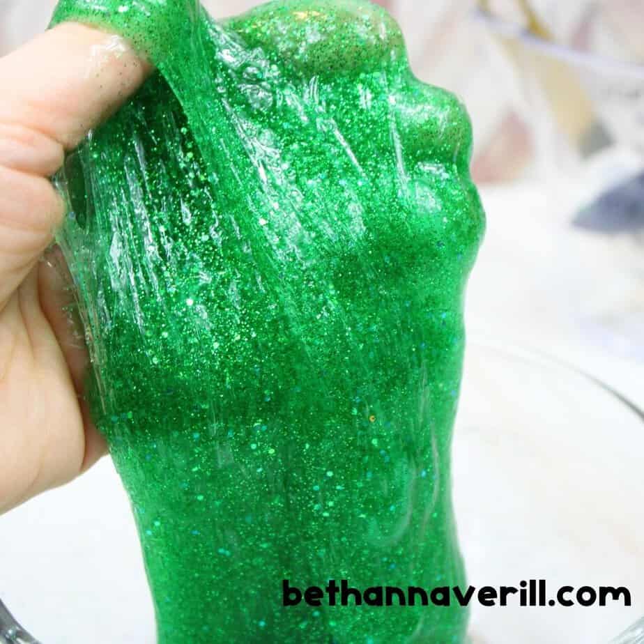 A hand stretches bright green glitter slime up and out of a bowl, showing its smooth, gooey, and elastic texture in close-up detail.